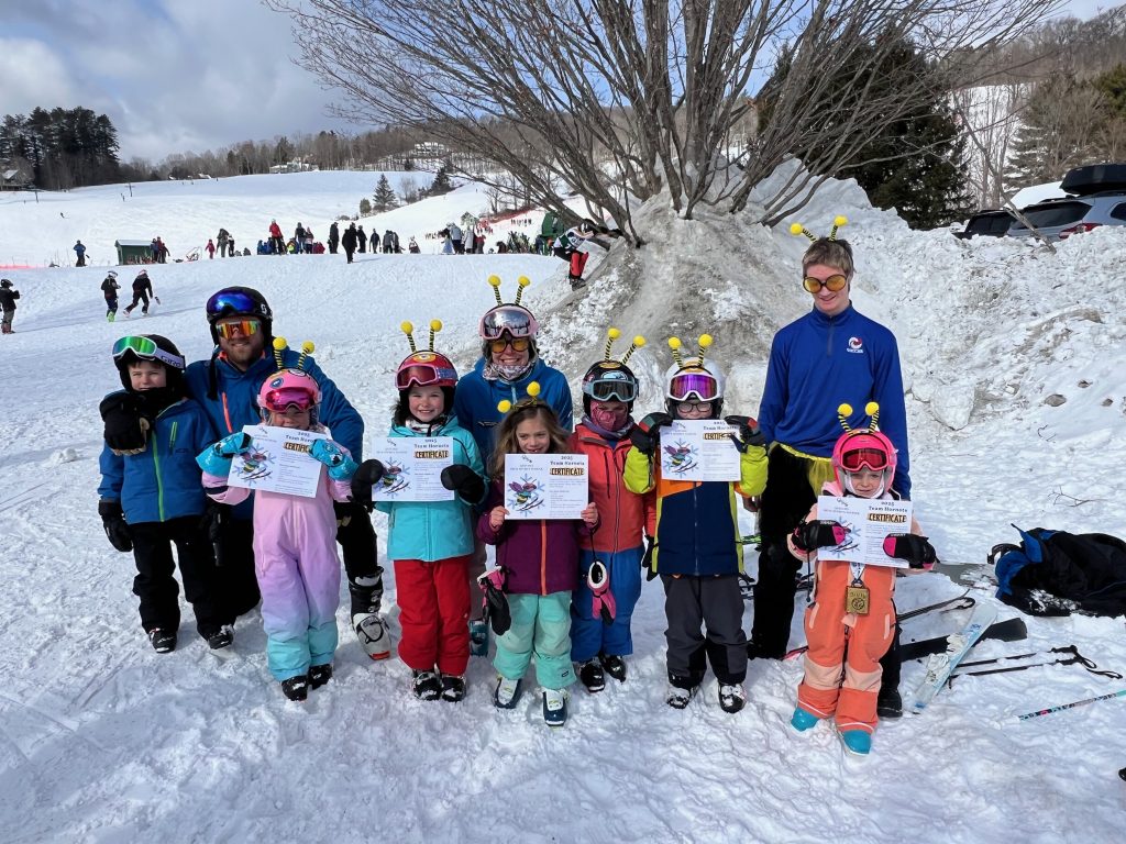 A group of young skiers and ski instructors wearing bee antennas and holding a certificate in the snow.