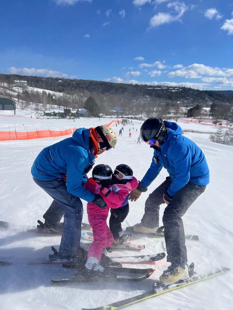 Two ski instructors in blue jackets at the top of the hill with two young skiers in pink jackets hugging each other.