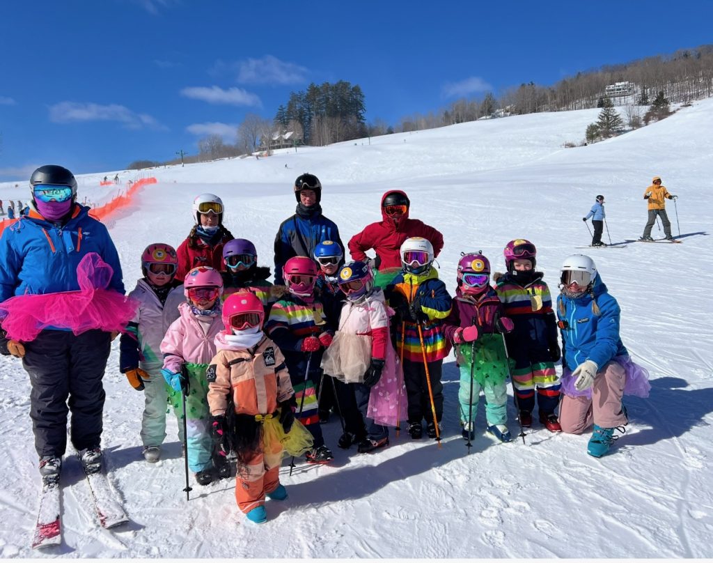 Ski instructors and skiers all wearing tutus on a sunny day with a blue sky behind them.